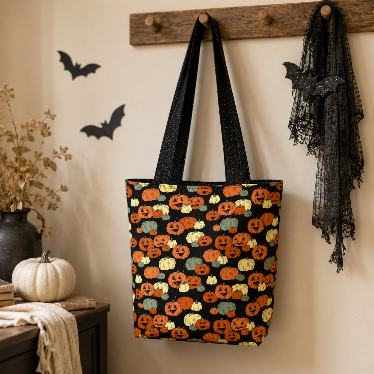 A black fabric tote bag with a colorful embroidered pumpkin pattern hangs from a wooden wall hook. The background is a cozy, decorated wall with Halloween elements including black bat cutouts, a black lace scarf, and a small white pumpkin on a table.